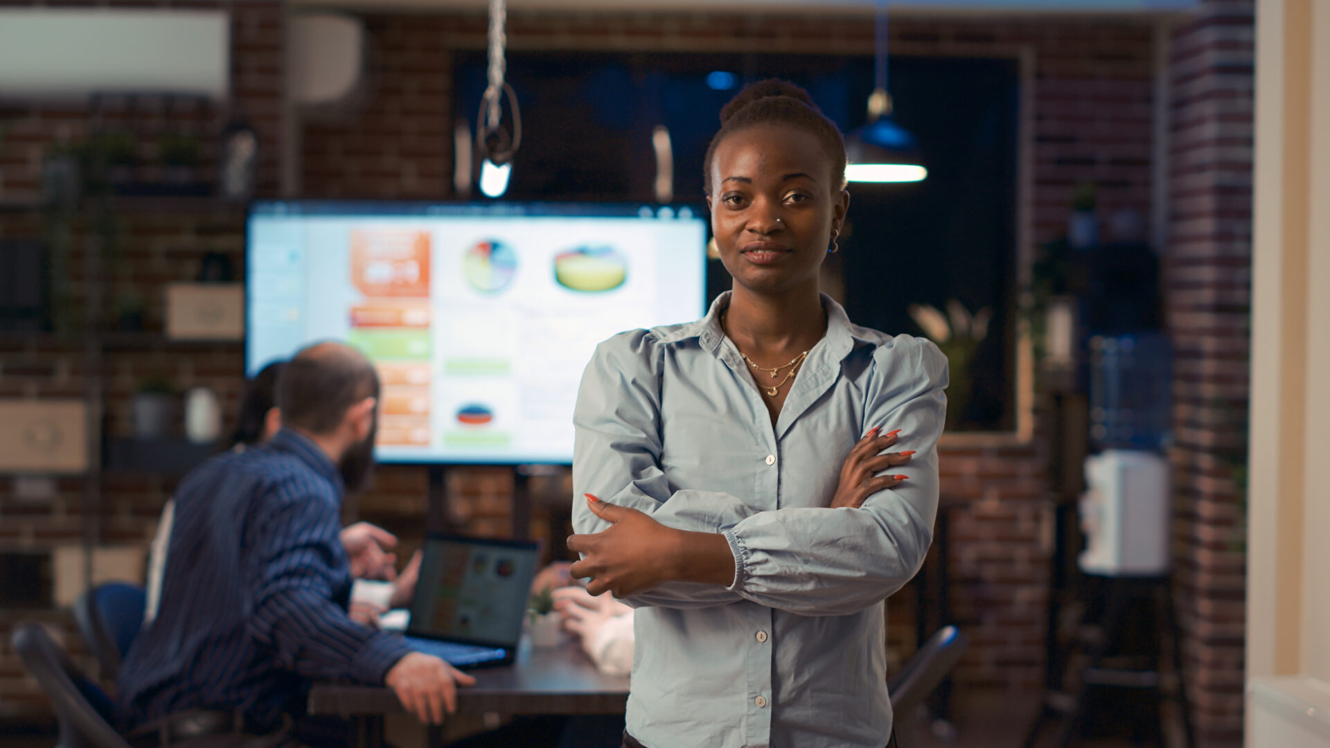 Smiling african american businesswoman crossing arms portrait, front view medium shot. Company employee standing in boardroom doorway in coworking space, coworkers presentation in background